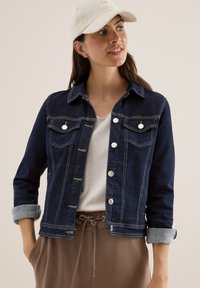 Mörk denimjacka med knäppning, två bröstfickor och uppvikta ärmar. Bärs över en ljus t-shirt, i kombination med avslappnade byxor.