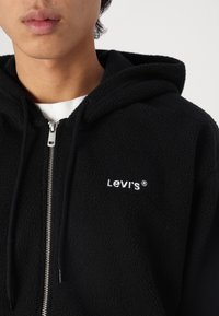 Ung person klädd i en svart fleece-hoodie från Levi's med en silvrig dragkedja som är delvis dragit upp över en vit skjorta, avskuren vid halsen och axlarna.
