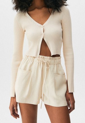 Kvinde iført beige ribbet cropped cardigan med to knapper og beige højttaljede shorts med snøre og store forlommer.
