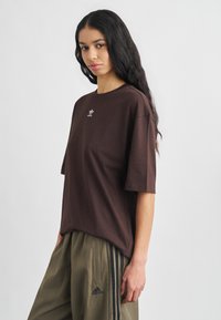 Νεαρή γυναίκα με μακριά σκούρα μαλλιά, φοράει καφέ μπλουζάκι Adidas και χακί παντελόνι Adidas με μαύρες ρίγες, στέκεται μπροστά σε μία απλή φόντο.