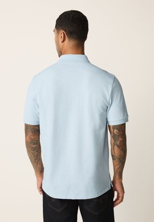 Mann mit Arm-Tattoos, der ein hellblaues kurzärmeliges Poloshirt und dunkle Jeans trägt, steht mit dem Rücken zu einem einfarbigen Hintergrund.