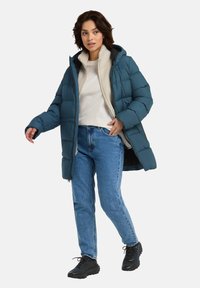 Giubbotto imbottito color teal con cappuccio, dotato di chiusura a zip, isolamento testurizzato e fodera interna morbida. Indossato sopra una camicia bianca e jeans.