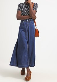 Falda maxi de mezclilla con aberturas laterales, cintura alta y dobladillo sin terminar. Combinada con una blusa ajustada y estampada y sandalias de plataforma marrones.