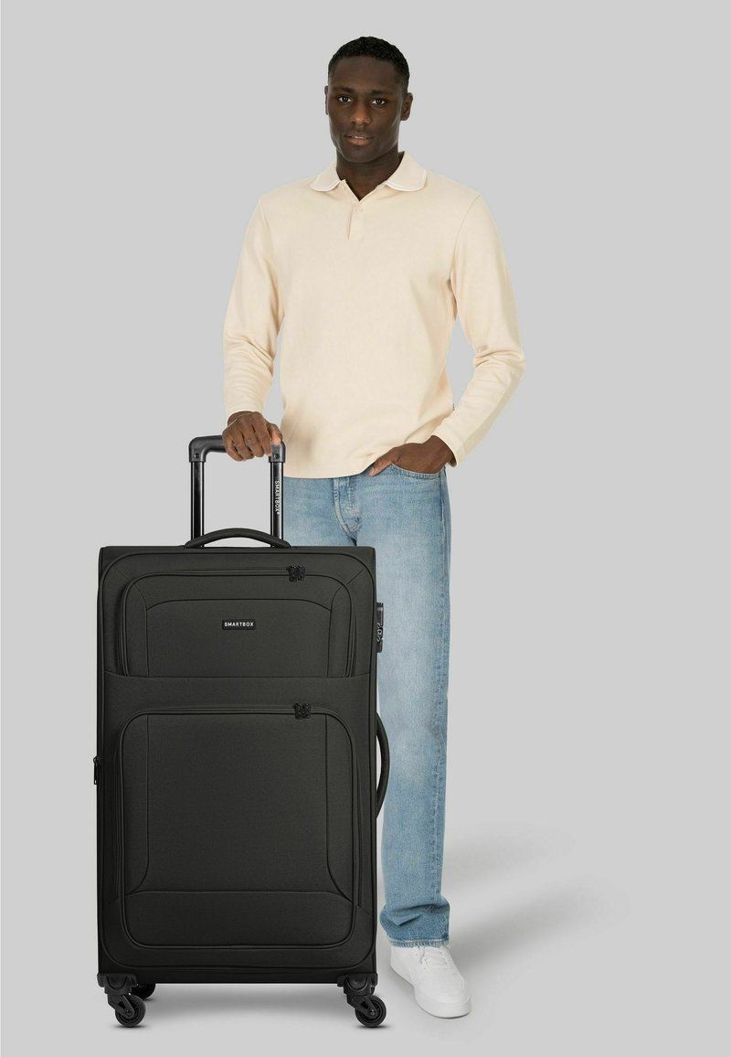 Hombre con camisa de manga larga beige y vaqueros, de pie sosteniendo el asa de una gran maleta negra con cuatro ruedas sobre fondo gris.