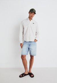 Camicetta a maniche lunghe a righe chiare, pantaloni corti in denim blu, sandali a scivolo marroni e un berretto verde scuro con logo. Abbigliamento semplice e casual.