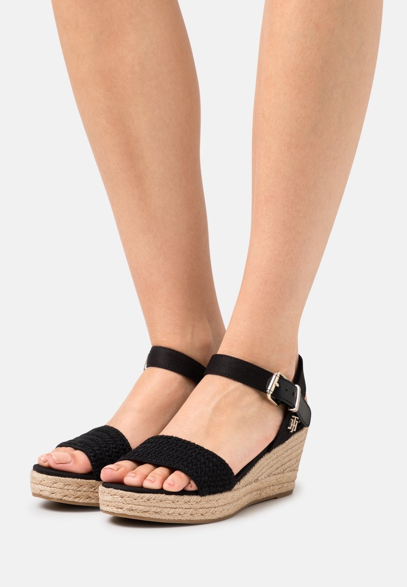 Pieds portant des sandales compensées noires en tissage avec brides arrière, des brides à la cheville et des talons compensés enveloppés de jute sur un fond uni.