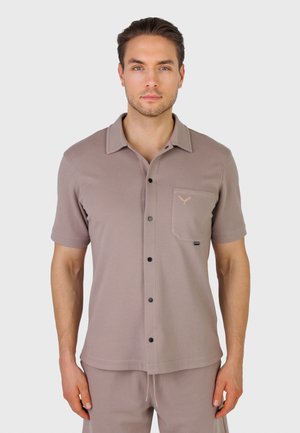 Mand iført taupe kortærmet skjorte med brystlomme og matchende shorts, stående foran en ensfarvet lys baggrund.