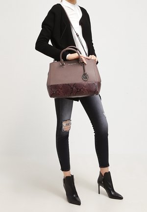 Mujer con cárdigan negro, camisa blanca, vaqueros pitillo negros desgarrados, botines negros de tacón, sosteniendo un bolso grande de cuero marrón con correa para el hombro.