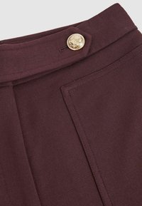 Közelkép egy bordó szövet derékpántjáról, díszes arany gombbal és részletes varrással a szabott ruházaton.