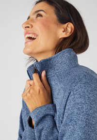 Chaqueta de forro polar azul, texturizada, con un alto cuello y cremallera. Primer plano de una mano sosteniendo el cuello, mostrando la suavidad de la tela.