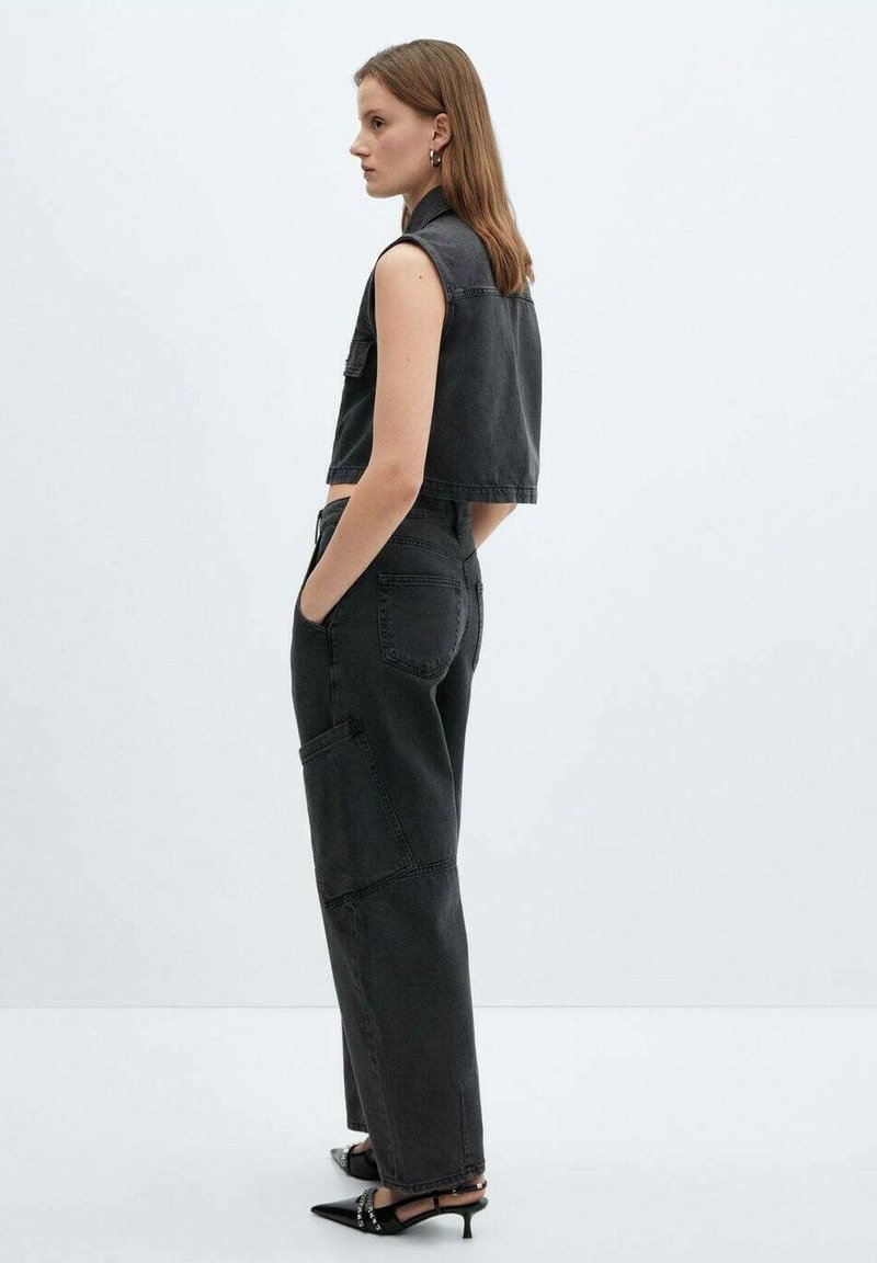 Gilet in denim nero e pantaloni a vita alta con tasche a toppa. Modella in piedi di lato, indossa scarpe con tacco nere con decorazioni.