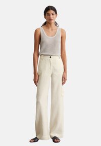 Leichtes, gestricktes beiges Tanktop kombiniert mit cremfarbigen, weit geschnittenen Hosen mit Taschen und einem Knopfverschluss. Schwarze flache Sandalen.