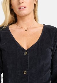 Schwarzes Cordtop mit V-Ausschnitt, das vorne drei Knöpfe und einen strukturierten Stoff aufweist. Betont durch eine zarte goldene Halskette.