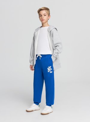 Szara bluza z zamkiem, biała koszulka, niebieskie spodnie dresowe z nadrukiem "NC" oraz białe sneakersy z zielonymi akcentami, wszystkie na neutralnym tle.