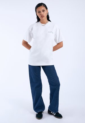 Weißes Baumwoll-T-Shirt mit kurzen Ärmeln, versehen mit einem kleinen grünen Logo. Kombiniert mit dunkelblauen Schlagjeans und schwarzen Sneakers mit weißen Streifen.