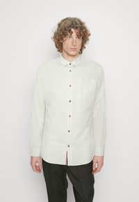 Camicia bianca a maniche lunghe realizzata in tessuto leggero, con chiusura a bottoni sul davanti, colletto a punta e una tasca sul petto con dettagli rossi e blu.