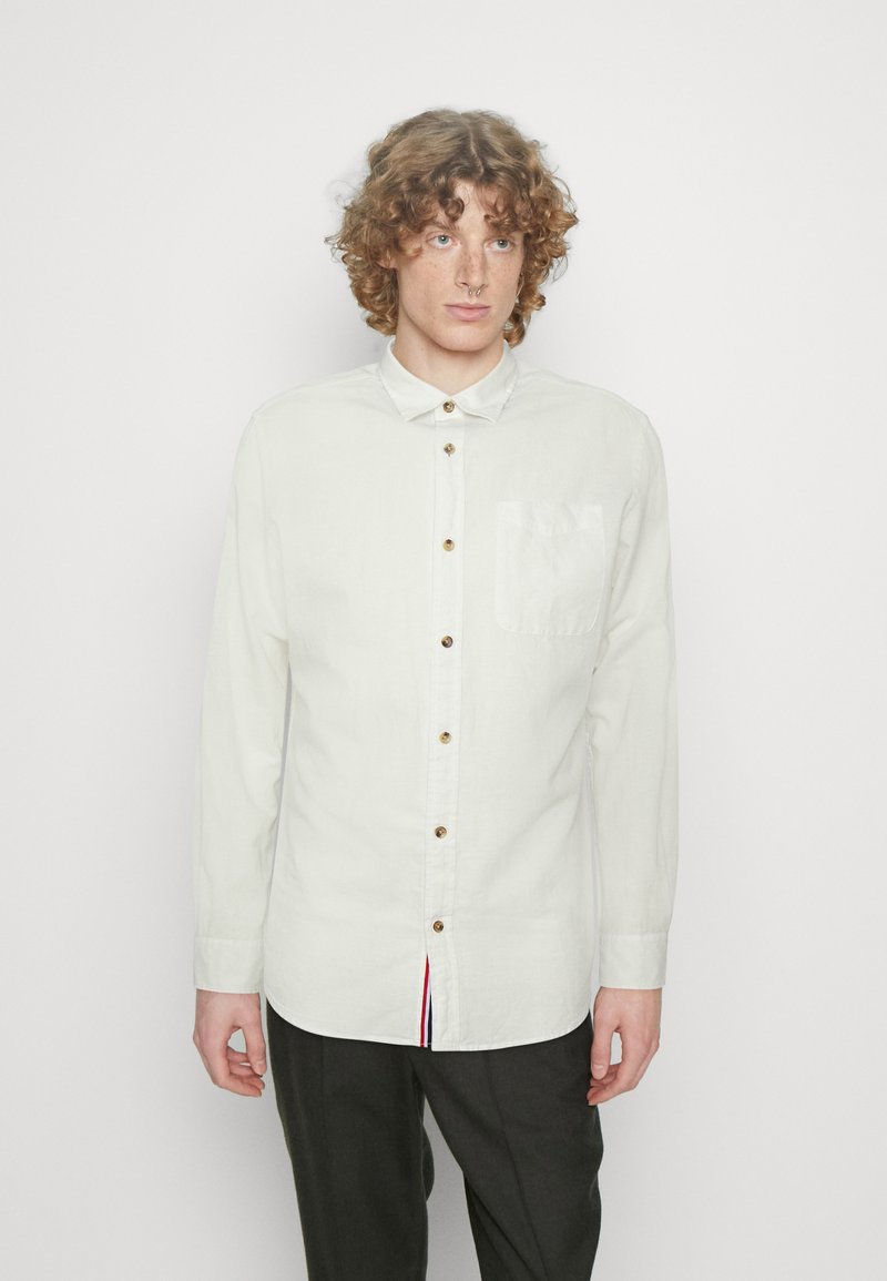Camicia bianca a maniche lunghe realizzata in tessuto leggero, con chiusura a bottoni sul davanti, colletto a punta e una tasca sul petto con dettagli rossi e blu.
