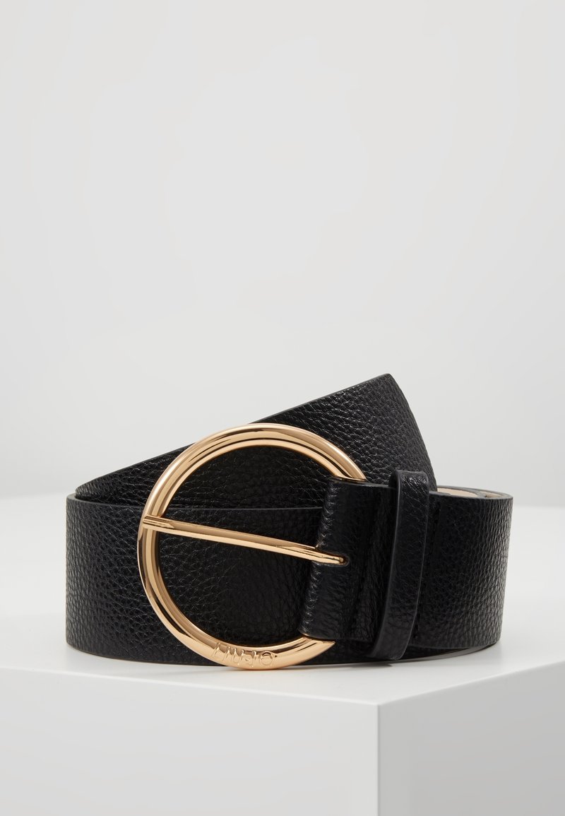 Ceinture en cuir noir texturé roulée sur une surface blanche avec une grande boucle ronde en métal doré.