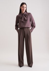 Braune gemusterte Bluse mit Schleife, die weiße Quadrate zeigt. Kombiniert mit hoch taillierten braunen Hosen und spitzen schwarzen Absätzen. Glattes Material.