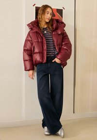 Bordó színű, kapucnis steppelt dzseki fényes anyagból, hozzá csíkos tengerészkék ing és sötét, bő szárú farmernadrág. Ezüst sportcipővel egészül ki az összeállítás.