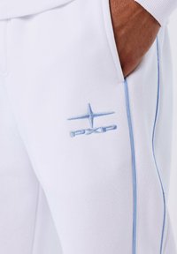 Close-up van de hand van een persoon in de zak van een witte broek met lichtblauwe piping en geborduurd blauw PXP-logo en ster.