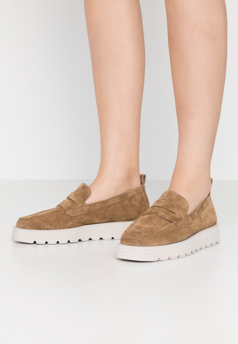 Pieds portant des mocassins en daim beige à enfiler avec des semelles épaisses blanches à rainures sur un fond blanc uni.