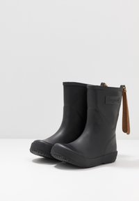 Des bottes noires en caoutchouc à la cheville avec une semelle texturée, un dessus lisse et une sangle en cuir marron avec un logo embossé pour un enfilage facile.