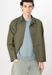 Olive green, lightweight jacket with a button closure and two front pockets, layered over a light blue sweatshirt and faded jeans.