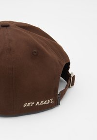 Brun bomull basebollkeps med kurvad skärm, justerbart spänne och den broderade frasen "FÖRBERED DIG" på baksidan.