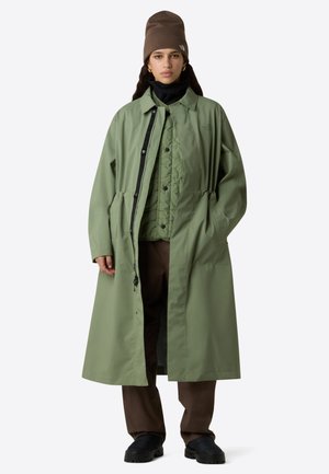 Cappotto impermeabile verde con vestibilità rilassata, chiusura frontale a pressione, vita elastica e grandi tasche. Indossato sopra uno strato interno trapuntato e pantaloni marroni.