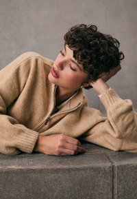 Beżowy sweter z wełny zapinany na zamek błyskawiczny z wysokim kołnierzem, ribbkim mankietami i gładką fakturą. Model opiera się na kamiennym podłożu, prezentując design odzieży.