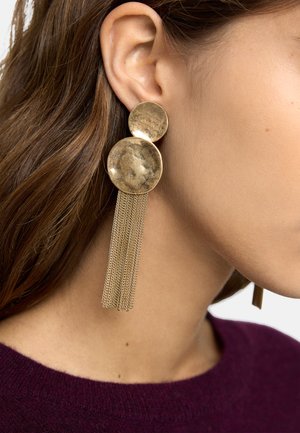 Złote kolczyki z podwójnymi dyskami i zwisającymi łańcuszkami noszone na lewym uchu, na tle jasnej skóry i brązowych włosów, w połączeniu z fioletową bluzką.