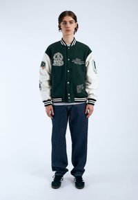 Giacca varsity verde e crema con toppe ricamate, colletto a coste e polsini a righe; abbinata a jeans in denim blu e sneaker nere.