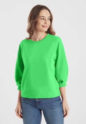 Vrouw met schouderlang haar, gekleed in een felgroene longsleeve en een blauwe spijkerbroek, die iets opzij kijkt en glimlacht.
