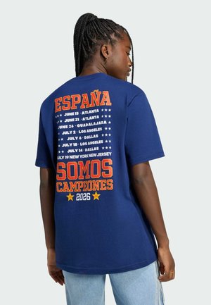 Femme aux cheveux tressés portant un t-shirt bleu avec les dates de la tournée espagnole et le texte "España Somos Campeones 2026" au dos.