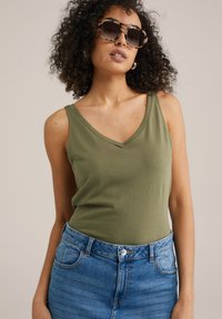 Olivno zelena brezrokavna majica s dekoltejem v obliki V, dopolnjena z modrimi jean kratkimi hlačami z visokim pasom. Model nosi oversize sončna očala.