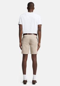 Beige bomuldsshorts med opretstående kant, parret med en almindelig hvid T-shirt og et brunt læderbælte. Udsigten bagfra viser minimal detaljering.