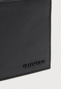 Crna teksturirana kožna površina sa zašivenim rubovima i crnim Valentino logotipom utisnutim u donjem desnom kutu.