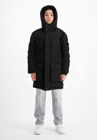 Zwarte gewatteerde parka met een capuchon, voorzien van twee voorzakken en drukknopen. Draagt over lichtblauwe versleten jeans en witte sneakers.