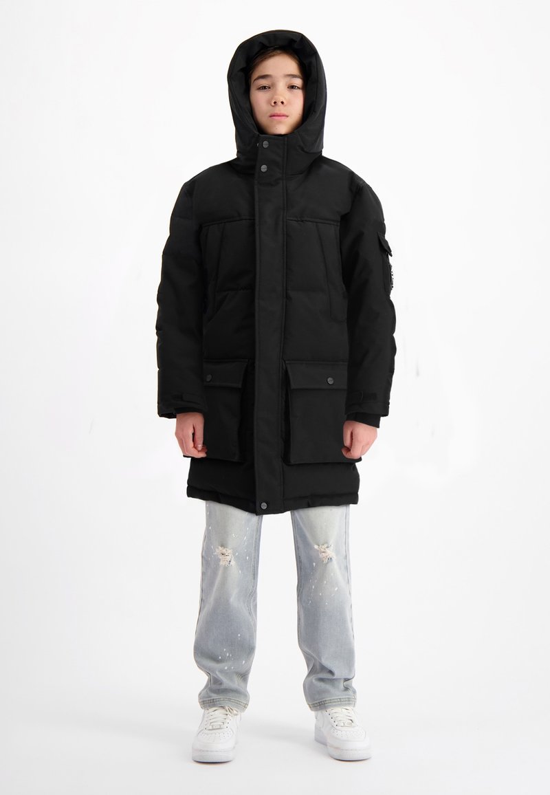 Zwarte gewatteerde parka met een capuchon, voorzien van twee voorzakken en drukknopen. Draagt over lichtblauwe versleten jeans en witte sneakers.