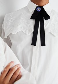 Hvit skjorte med en blond, brodert bærestykke og et svart satengslips prydet med en blå og sølv juvel ved kragen. Glatt stoffstruktur.