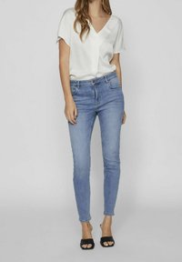 Top blanco de cuello en V, jeans ajustados azul claro y sandalias de tacón negras. El top tiene mangas relajadas; los jeans tienen bolsillos frontales y cierre con botón.