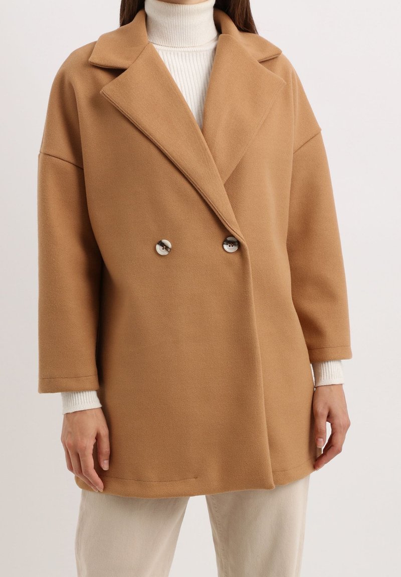 Abrigo de color camello de doble botonadura con solapa, grandes botones y silueta relajada. Presenta mangas anchas y una textura suave.