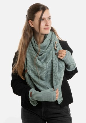Jonge vrouw met lang haar, gekleed in een zwarte trui, een lichtblauwe gebreide sjaal en bijpassende zondervingerhandschoenen, die naar de zijkant kijkt.