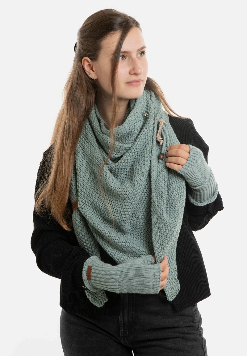 Jonge vrouw met lang haar, gekleed in een zwarte trui, een lichtblauwe gebreide sjaal en bijpassende zondervingerhandschoenen, die naar de zijkant kijkt.