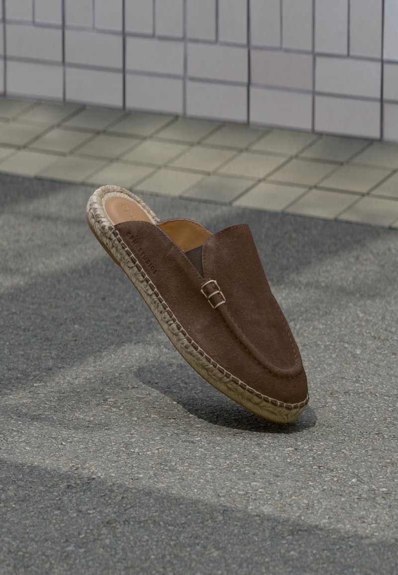 Καφέ σουέντ μοκασίνι χωρίς κορδόνια με πλεκτή σόλα από τζούτα να αιωρείται πάνω από άσφαλτο κοντά σε πλακιδωμένο τοίχο.