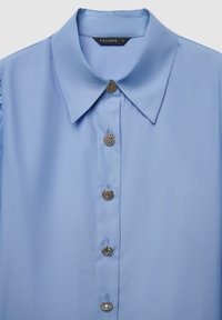 Light blue shirt with a structured collar, featuring five decorative buttons, including round and knot designs, made of smooth fabric.
