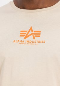 Beige puuvillane t-särk ümmarguse kaelusega ja oranž "Alpha Industries" logo, millel on stiliseeritud kiri ja graafilised elemendid.