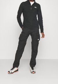 Svart fleece-tröja med kvartsgång, svarta cargobyxor med flera fickor och robusta flerfärgade sneakers med en design i vitt, svart och orange.