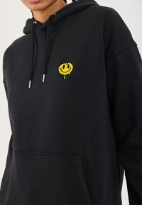Sweat à capuche noir en coton avec une poche kangourou à l'avant. Présente un visage souriant jaune brodé sur la poitrine avec des détails dégoulinants.